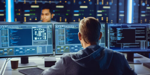 Person working at a desk with multiple monitors displaying code and data, in a secure operations or data center environment
