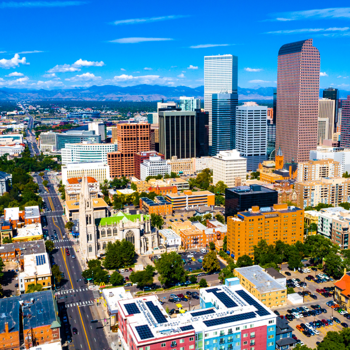 A sunny, aerial view of downtown Denver, Colorado, featuring modern skyscrapers, historic buildings, and tree-lined streets, with the Rocky Mountains in the background—highlighting a hub for temp staffing and consulting.