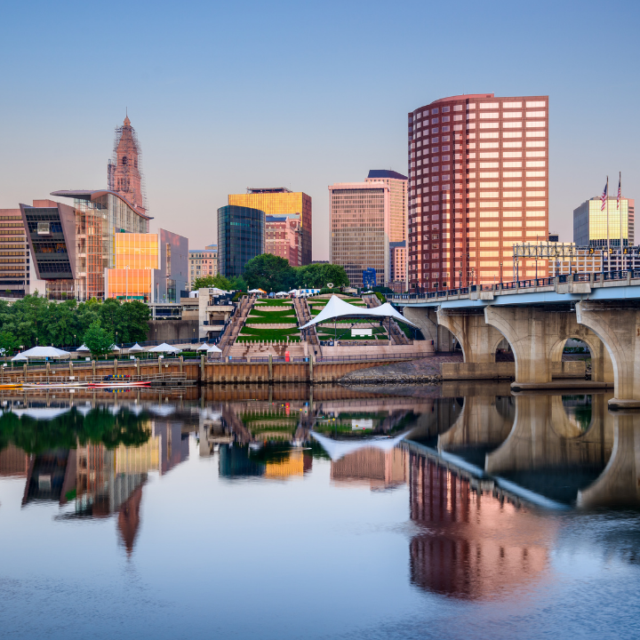 A serene view of Hartford, Connecticut’s skyline at dusk, with modern glass buildings, a pedestrian-friendly riverfront, and a stone bridge reflecting in the calm water—representing nearby Glastonbury's access to top temp agency services.