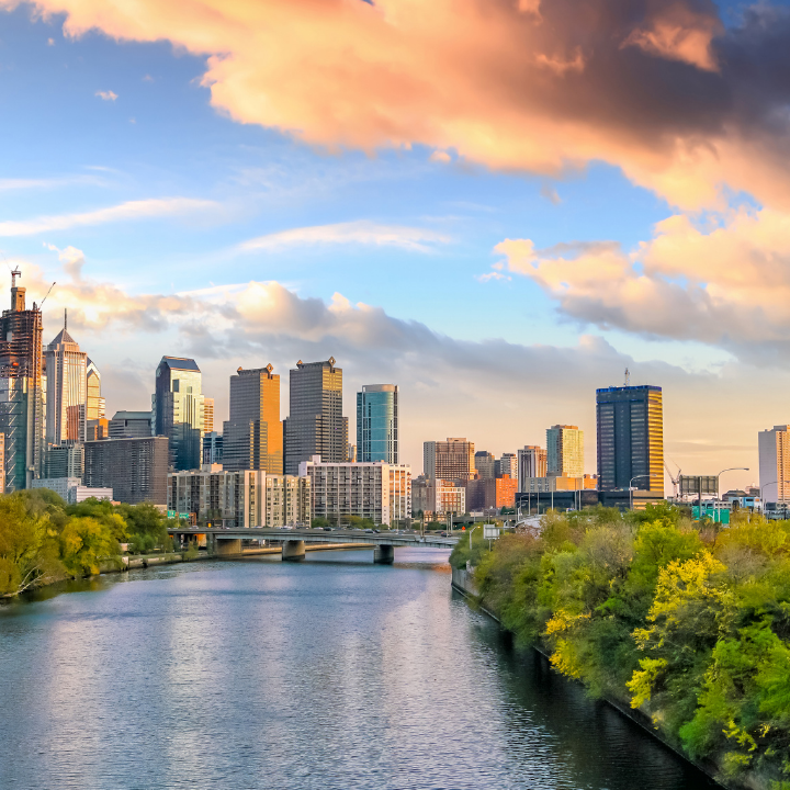 A scenic view of downtown Philadelphia with high-rise buildings reflecting the evening sun, the Schuylkill River flowing in the foreground, and lush trees lining the water—symbolizing Philly’s thriving temp staffing and consulting scene.