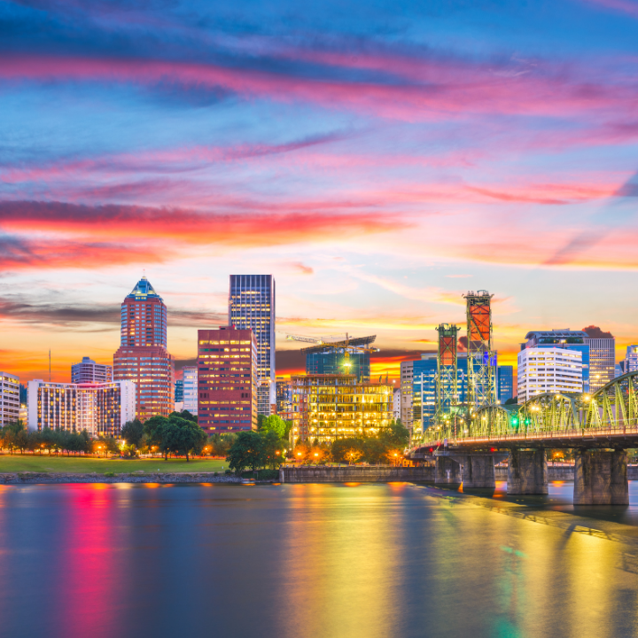 A vibrant sunset over downtown Portland, Oregon, with illuminated high-rises reflecting in the Willamette River and a prominent bridge in the foreground—representing the region served by trusted Oregon recruiters.