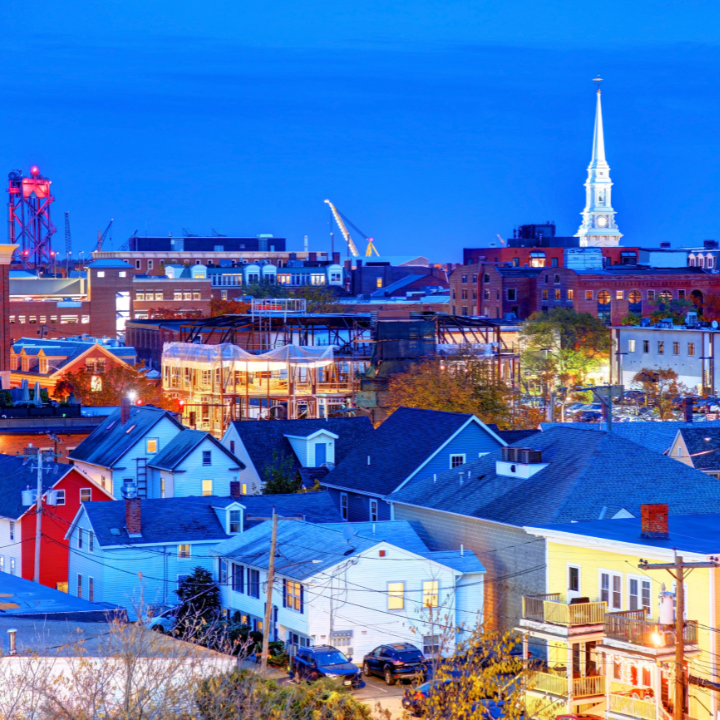 An evening view of Portsmouth, NH, highlighting colorful homes, historic brick buildings, a glowing construction site, and a white church steeple—capturing the city where staffing and consulting thrive in a vibrant coastal community.