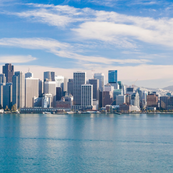 A bright, clear view of downtown San Francisco’s skyline from across the bay, featuring modern high-rises, calm blue waters, and a bustling business district—representing the city served by temp staffing and consulting experts.
